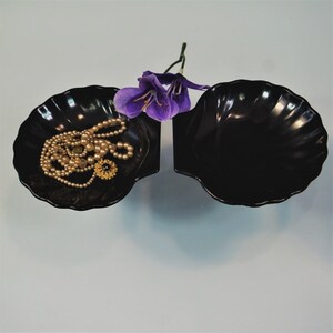 May include: Two black shell-shaped dishes with scalloped edges. One dish contains a multi-strand cream-colored pearl necklace with a gold-toned floral pendant. A single purple artificial flower rests between the two dishes.