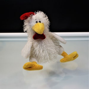 May include: A plush white chicken toy with a red comb and bow tie, yellow beak and feet. The toy has a fluffy texture and is set against a white and black background.