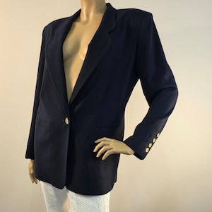 May include: A navy blue blazer featuring structured shoulders, a single gold-tone button closure at the front, and three smaller matching gold-tone buttons on each sleeve cuff. The garment has a notched lapel collar and two front welt pockets.