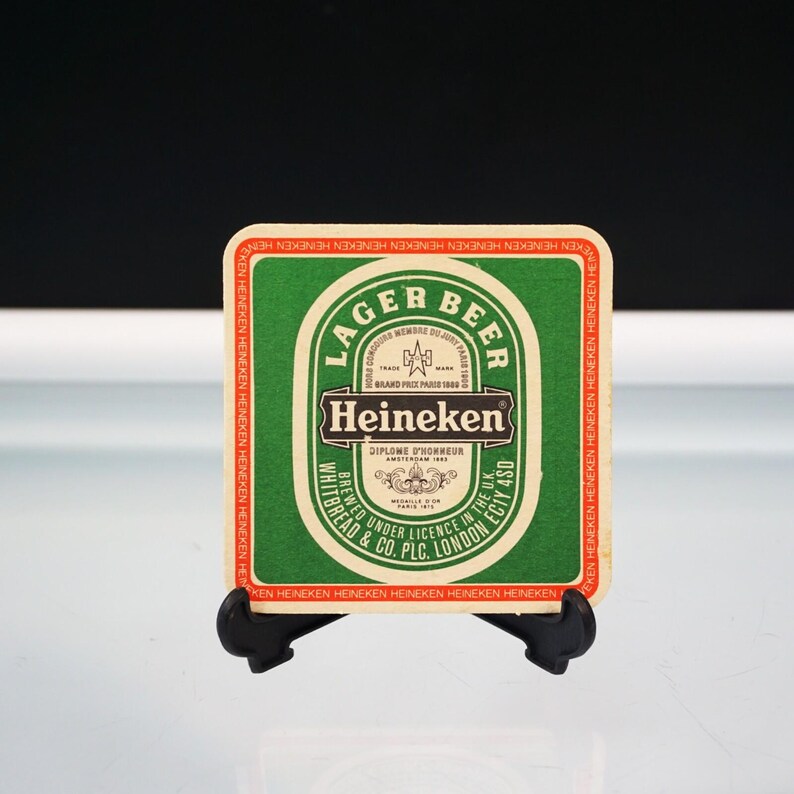 Puede incluir: Un posavasos cuadrado verde y blanco de cerveza Heineken. El posavasos presenta el logotipo y el texto de Heineken, incluyendo "Lager Beer" y "Brewed Under Licence in the U.K.". El posavasos se muestra en un soporte negro.