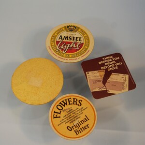 Puede incluir: Se muestran cuatro posavasos. Uno es redondo con el texto "AMSTEL Light" en rojo y blanco. Otro posavasos redondo tiene el texto "FLOWERS Original Bitter". Un tercer posavasos es un c&iacute;rculo amarillo liso. El cuarto posavasos es un cuadrado marr&oacute;n con el texto "THINK BEFORE YOU DRINK BEFORE YOU DRIVE".