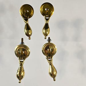 May include: Four vintage brass drawer pulls. Each pull features a circular backplate, a decorative handle, and a teardrop-shaped pull. The brass hardware is in a polished finish, with visible screws and nuts for mounting.