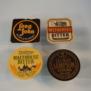 May include: Four vintage drinks coasters. A black square coaster with "Long John SCOTCH WHISKY" in yellow and orange. A beige, octagonal coaster with "WETHERED'S BITTER" in red. A yellow circular coaster with "Badger MALTHOUSE BITTER". A black circular coaster with "TRUMAN SAMPSON STRONG BITTER".