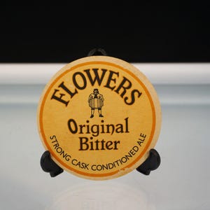 Puede incluir: Un posavasos redondo y beige con el texto "FLOWERS Original Bitter" en negro. Una peque&ntilde;a ilustraci&oacute;n de un hombre con un barril est&aacute; en el centro. Las palabras "STRONG CASK CONDITIONED ALE" est&aacute;n en el borde.