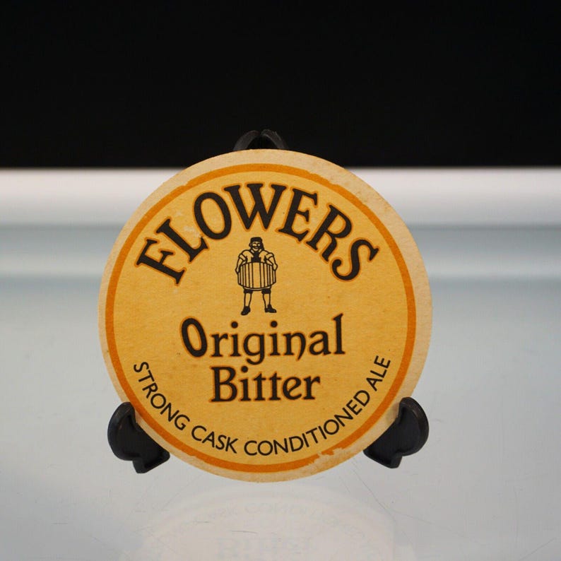 Puede incluir: Un posavasos redondo y beige con el texto "FLOWERS Original Bitter" en negro. Una peque&ntilde;a ilustraci&oacute;n de un hombre con un barril est&aacute; en el centro. Las palabras "STRONG CASK CONDITIONED ALE" est&aacute;n en el borde.