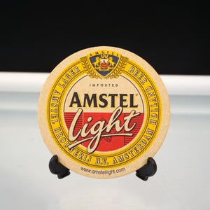 Puede incluir: Posavasos redondo de cerveza Amstel Light. El posavasos es de color beige con texto rojo y blanco que dice "AMSTEL Light". La parte superior del posavasos tiene un emblema dorado y azul. La parte inferior del posavasos tiene el texto "BROUWERIJ B.V. AMSTERDAM".