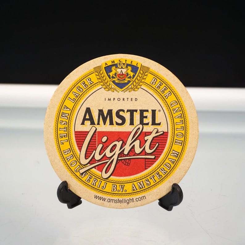 Puede incluir: Posavasos redondo de cerveza Amstel Light. El posavasos es de color beige con texto rojo y blanco que dice "AMSTEL Light". La parte superior del posavasos tiene un emblema dorado y azul. La parte inferior del posavasos tiene el texto "BROUWERIJ B.V. AMSTERDAM".