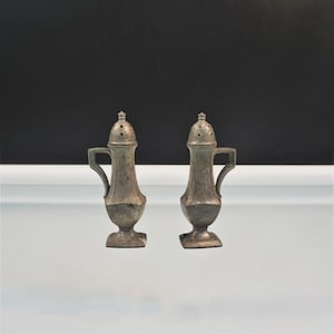 May include: A pair of matching metallic salt and pepper shakers with a weathered gray finish. Each shaker features a tall, slender body with a square base, a curved handle on one side, and a rounded, perforated top for dispensing seasonings.
