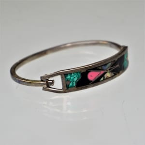 May include: Silver-toned hinged bangle bracelet with a decorative rectangular panel. The panel features a black background with inlaid turquoise, pink, and iridescent shell-like accents. The bracelet is closed with a simple clasp.