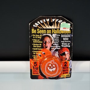 May include: A Safety Strobe light in orange packaging with a pumpkin graphic. The card features text including Be Seen on Halloween, 5 Year Limited Warranty, and 2 Watch Type Batteries Included, alongside images of children in costumes.