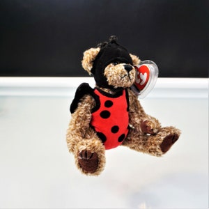 May include: A brown plush teddy bear wearing a black headpiece and a red vest with black polka dots. The bear has dark brown paw pads and a clear plastic heart-shaped tag attached to its ear with the red and white text ty printed on the front.
