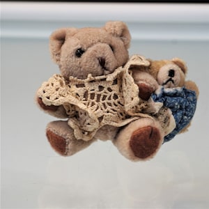 May include: Two plush teddy bears. The larger bear is light brown and wears a cream-colored crocheted garment. A smaller bear, with a blue patterned garment, is nestled beside it. Both bears have dark brown eyes and noses.