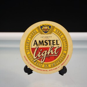 Puede incluir: Un posavasos redondo de cerveza Amstel Light. El posavasos tiene un fondo beige con texto amarillo y rojo. El texto incluye "AMSTEL Light", "IMPORTED" y "BEER". El posavasos est&aacute; sobre un soporte negro.