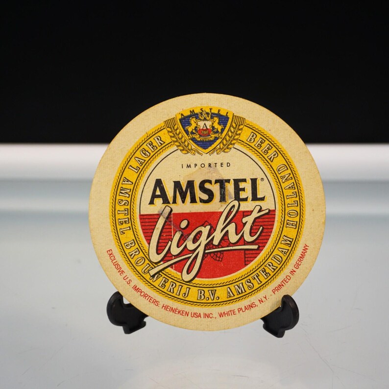 Puede incluir: Un posavasos redondo de cerveza Amstel Light. El posavasos tiene un fondo beige con texto amarillo y rojo. El texto incluye "AMSTEL Light", "IMPORTED" y "BEER". El posavasos est&aacute; sobre un soporte negro.