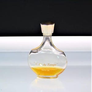 May include: A clear glass perfume bottle with a silver-colored cap. The bottle contains a yellow liquid and has the words "L'Air du Temps" and "Bath Oil" printed on it. The bottle is sitting on a white surface.