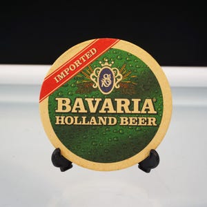 Puede incluir: Un posavasos redondo con un dise&ntilde;o verde y beige. El posavasos presenta el texto "Bavaria Holland Beer" e "Imported". Una banda roja contiene la palabra "Imported".
