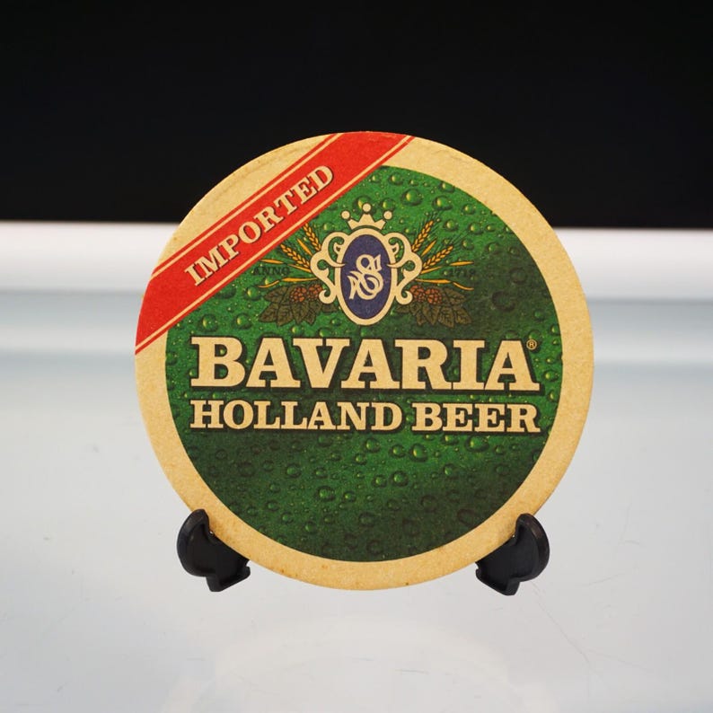 Puede incluir: Un posavasos redondo con un dise&ntilde;o verde y beige. El posavasos presenta el texto "Bavaria Holland Beer" e "Imported". Una banda roja contiene la palabra "Imported".