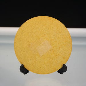 Puede incluir: Un posavasos redondo y amarillo con un parche cuadrado de color m&aacute;s claro en el centro. Se muestra sobre un soporte negro sobre un fondo blanco y negro. El posavasos est&aacute; hecho de un material texturizado.