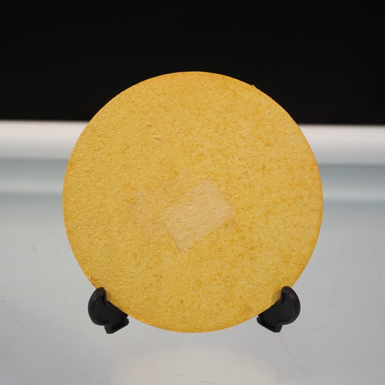 Puede incluir: Un posavasos redondo y amarillo con un parche cuadrado de color m&aacute;s claro en el centro. Se muestra sobre un soporte negro sobre un fondo blanco y negro. El posavasos est&aacute; hecho de un material texturizado.