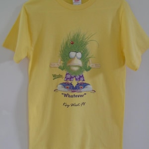 May include: Yellow t-shirt featuring a cartoon character with green hair, a red ladybug, and blue shoes. The shirt has the text "Whatever" and "Key West, FL".
