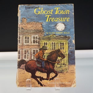 May include: Vintage paperback book titled "Ghost Town Treasure" by Clyde Robert Bulla. The cover illustration depicts a cowboy on horseback riding through a ghost town under a moonlit sky. Illustrated by Robert Shore.