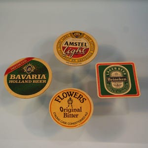 Puede incluir: Cuatro posavasos de cerveza con varios logotipos de marcas. Incluye un posavasos redondo con "Amstel Light", un posavasos verde y dorado "Bavaria Holland Beer", un posavasos cuadrado "Heineken" y un posavasos redondo "Flowers Original Bitter".