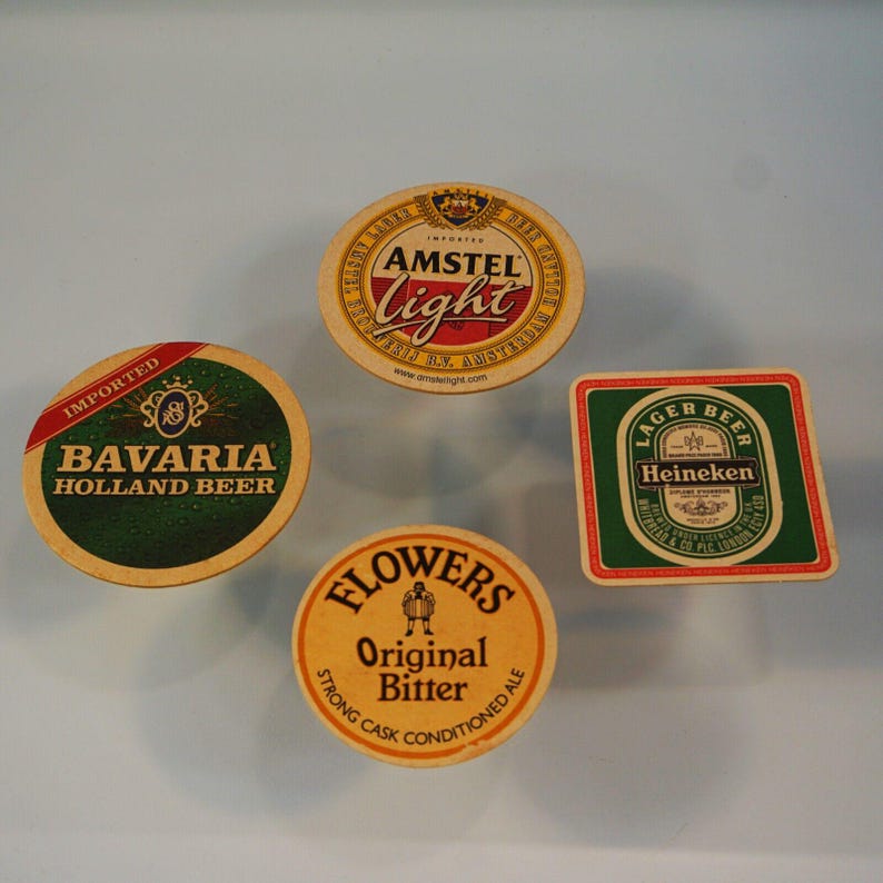 Puede incluir: Cuatro posavasos de cerveza con varios logotipos de marcas. Incluye un posavasos redondo con "Amstel Light", un posavasos verde y dorado "Bavaria Holland Beer", un posavasos cuadrado "Heineken" y un posavasos redondo "Flowers Original Bitter".