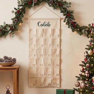 May include: A hanging fabric advent calendar in off-white canvas featuring the name Caleb in dark green script at the top. The calendar has twenty-four individual pockets arranged in six rows, each printed with a gold number from one to twenty-four.