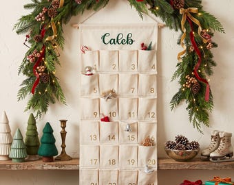 Può includere: Un calendario dell'avvento in tessuto color crema appeso a un bastone di legno, con ventiquattro tasche numerate in oro. Il nome Caleb è ricamato in verde nella parte superiore. Il calendario è esposto sotto una ghirlanda sempreverde.