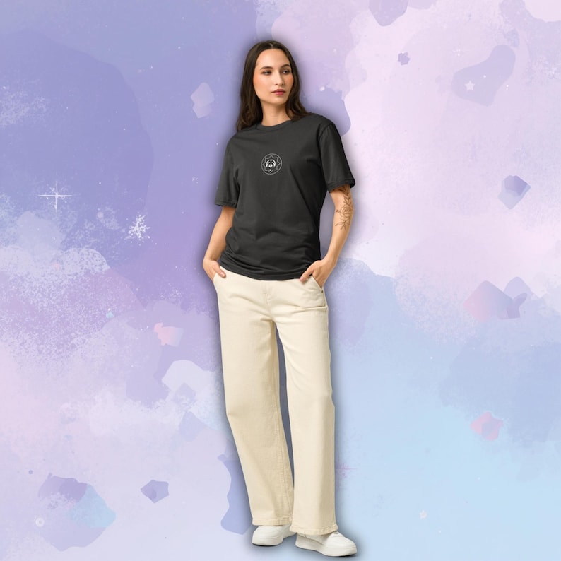 May include: A person wearing a black t-shirt with a circular graphic and cream-colored pants. The t-shirt has a small, white graphic on the chest. The pants are wide-legged and paired with white shoes. The background is a watercolor wash of purple and blue.