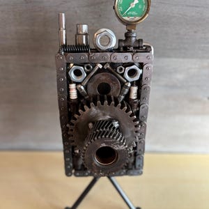 May include: A unique sculpture crafted from repurposed metal components, including gears, chains, and bolts. The artwork features a green gauge and is mounted on a tripod base. The industrial-style piece showcases a creative use of mechanical parts.