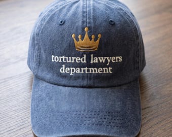 May include: A blue denim baseball cap featuring an embroidered gold crown above the white text that reads tortured lawyers department. The cap has a structured front panel, a curved brim, and stitched eyelets for ventilation on the crown.