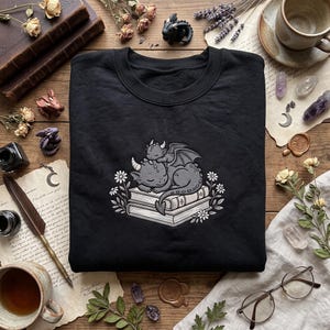 May include: A black crewneck sweatshirt featuring a graphic print of two grey dragons sleeping on a stack of books, surrounded by small white flowers. The design is centered on the chest of the garment, showcasing a detailed illustration of the creatures.