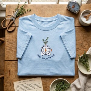 May include: A light blue t-shirt featuring an embroidered graphic of a kitchen timer with a sprig of thyme emerging from the top. Below the timer, the text reads "TAKE YOUR THYME" in dark blue letters. The shirt has short sleeves and a crew neckline.