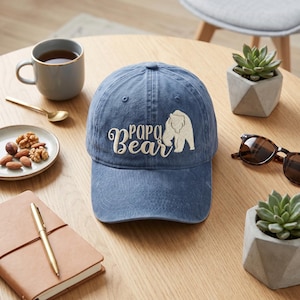 May include: A blue baseball cap with the embroidered text "Papa Bear" and a bear graphic. The hat is on a wooden table with a cup of tea, a notebook, and a small succulent plant.