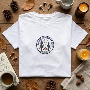 May include: White t-shirt with a circular embroidered design featuring trees, a mushroom, a crescent moon, and the text "FOREST INTROVERT CLUB". The shirt is displayed on a wooden surface with autumn-themed decorations.