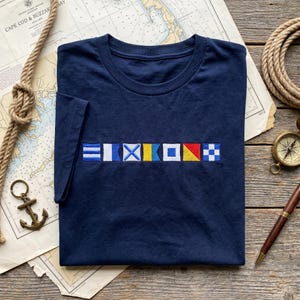 May include: A navy blue short-sleeve t-shirt featuring an embroidered row of seven colorful nautical signal flags across the chest. The flags include various geometric patterns in blue, white, yellow, and red, representing a maritime code.