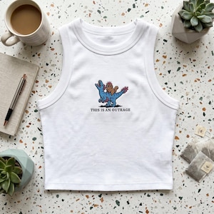 May include: A white ribbed sleeveless tank top featuring a central embroidered graphic of a blue dinosaur with a small brown creature wearing a crown on its back, accompanied by the black text "THIS IS AN OUTRAGE" printed below the design.