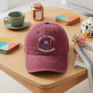 May include: A maroon baseball cap featuring a graphic of a green frog wearing a purple wizard hat and robe, holding a staff. The text "OFF TO COMMIT SHENANIGANS" is printed in white in an arc around the frog illustration on the front panel.