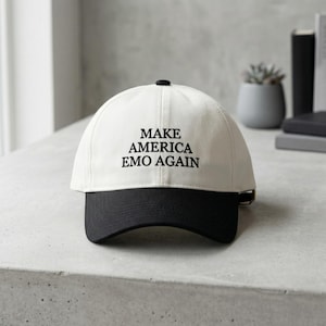 May include: A white baseball cap with a black brim. The text "MAKE AMERICA EMO AGAIN" is printed in black on the front of the cap. The cap is sitting on a light gray surface.