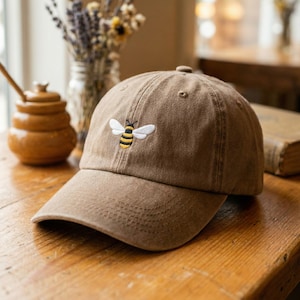 May include: A brown baseball cap with a curved peak. The cap has an embroidered bee design with yellow and black stripes and white wings. The cap is made of a soft material and is suitable for casual wear.