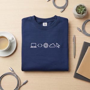 May include: Navy blue sweatshirt with white graphic icons representing coding and technology. The icons include a laptop, angle brackets, a gear, a cloud, and a mouse cursor. The sweatshirt is folded neatly on a light wood surface.