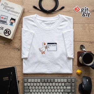 May include: A white short-sleeved t-shirt featuring an embroidered graphic of a goose standing next to a computer window that displays the text "hello, world!" with a blinking cursor. The design is centred on the chest of the garment.