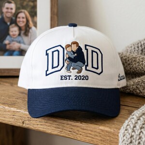 May include: White and navy blue baseball cap with the word "DAD" embroidered in navy blue block letters. The cap features an embroidered image of a father and child, with "EST. 2020" below. The name "Lucas" is embroidered on the side.