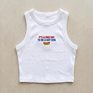 May include: A white ribbed tank top featuring an embroidered graphic on the chest. The text reads IT'S A BAD DAY TO BE A HOT DOG in red and blue lettering, positioned above a small, detailed embroidered illustration of a hot dog in a bun.