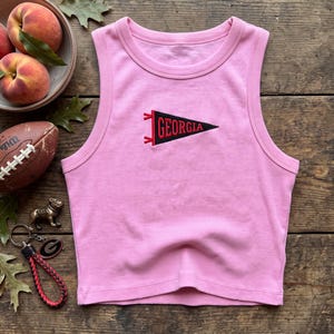 May include: A light pink ribbed tank top featuring a centered embroidered black pennant patch with the red text GEORGIA. The sleeveless garment has a round neckline and a straight hem, showcasing a simple design with a textured fabric finish.