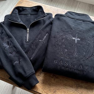 May include: Two black quarter-zip sweatshirts. One features a silver zipper, a sword, and bird embroidery on the sleeves. The other displays a large embroidered dragon, laurel wreath, and the text Basgiath War College on the back.