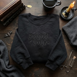 May include: A black crewneck sweatshirt featuring black embroidery on the chest that reads Surgical Tech in a gothic font, accompanied by small embroidered bat silhouettes and a scalpel icon. The garment has long sleeves and ribbed cuffs.
