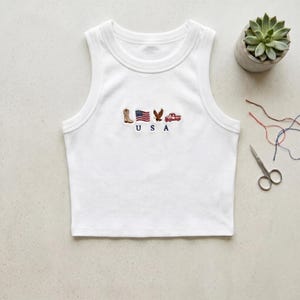 May include: A white ribbed tank top featuring a small embroidered design on the chest. The embroidery includes a cowboy boot, an American flag, an eagle, and a red truck, with the letters U S A stitched in navy blue thread below the icons.