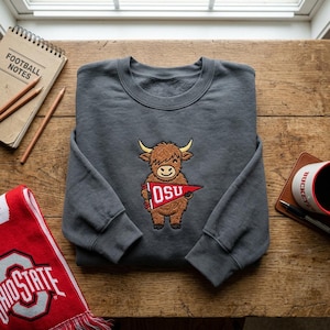 May include: A charcoal grey crewneck sweatshirt featuring an embroidered brown Highland cow holding a red pennant with the white text OSU. The sweatshirt is displayed with a notebook, two pencils, a red scarf, and a mug on a wooden surface.